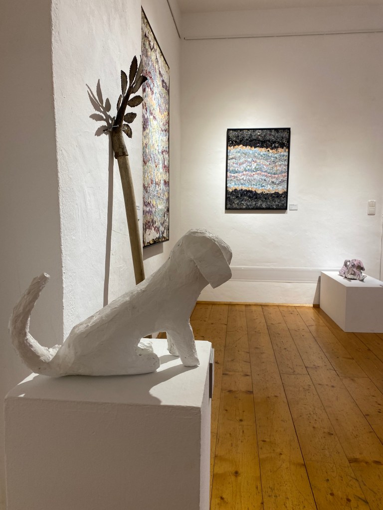 A white sculpture of a dog on a pedestal in an art gallery, surrounded by abstract paintings and wooden flooring. Work of art by florian Nörl, Textilstein 